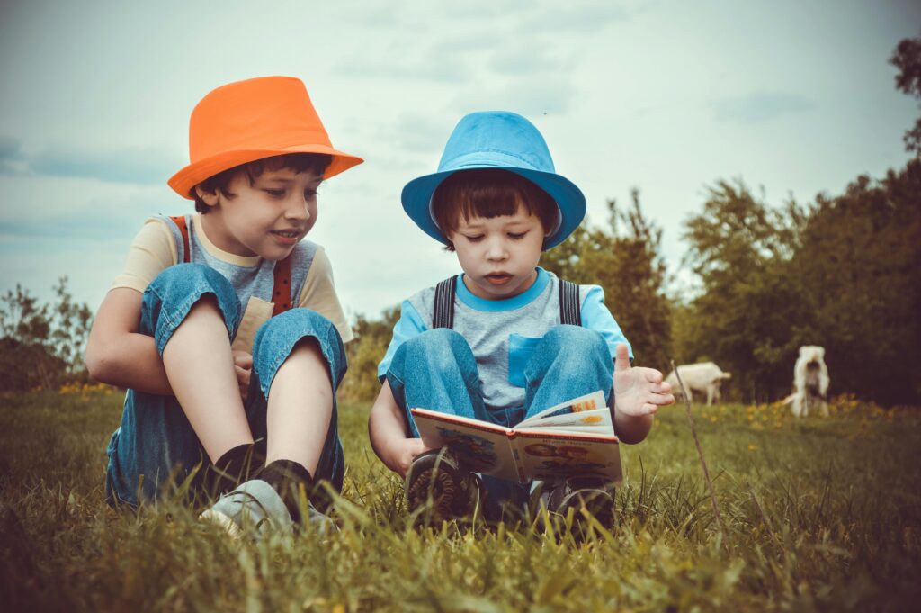 pexels photo 1094072 1094072 Two young boys enjoying a book together outdoors on a sunny day, wearing vibrant hats.