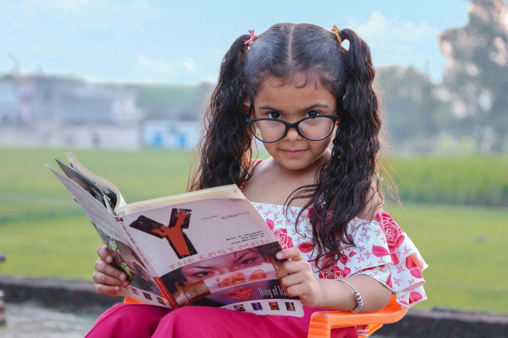 pexels photo 4191698 4191698 Funny ethnic little girl with curly pigtails and eyeglasses sitting on chair in park and reading magazine with interest